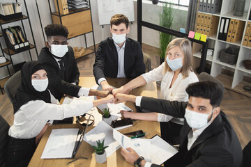 Five multi ethnic company workers in face masks and formal clothes fist bumping while having meeting during pandemic. Business colleagues sitting at desk and looking at camera.