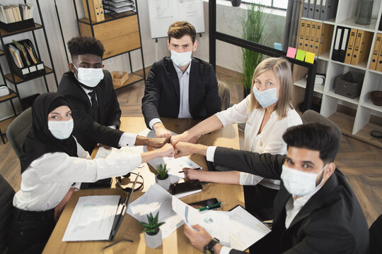 Five multi ethnic company workers in face masks and formal clothes fist bumping while having meeting during pandemic. Business colleagues sitting at desk and looking at camera.
