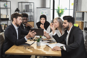 Group of five multi ethnic financial experts dressed in formal clothes sitting together at office desk and looking on tablet screen. Male and female colleagues using modern gadget during conference.