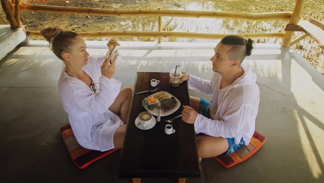 Attractive young man and woman eat and drink tasty beverages spending time in traditional local floating cafe on water. Loving happy couple having breakfast outdoors, feed each other. Tropical view
