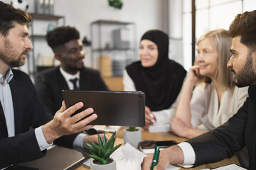 Multi ethnic team of business partners looking on one tablet screen during brainstorming at conference room. Five people discussing common working project at office.