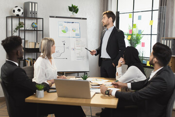 Confident caucasian businessman making presentation on flip chart for his multiracial colleagues that sitting together at desk. Group of successful business people having negotiation at boardroom.
