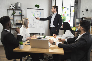 Successful man in black suit standing near flip chart and talking with multicultural colleagues during conference meeting. Group of business people gathering at office room for briefing.
