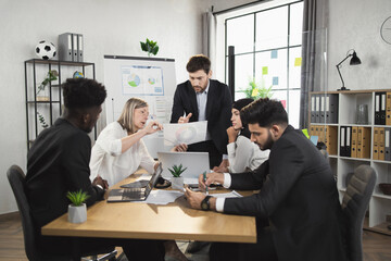 Diverse group of business partners discussing development of common project during meeting at boardroom. Colleagues doing paperwork and using modern technology.