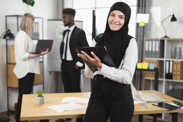 Muslim businesswoman with clipboard in hands smiling and looking at camera while her multiracial colleagues talking on background. Meeting at office of successful partners.