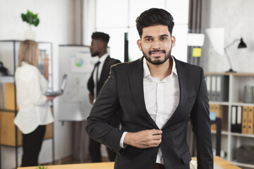 Successful indian man in suit smiling and looking at camera while his multi ethnic colleagues talking on background. Office room for negotiation.