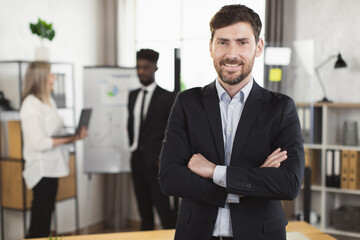 Portrait of positive caucasian businessman in suit looking and smiling on camera at boardroom while his multi ethnic colleagues talking on background. People and cooperation concept.