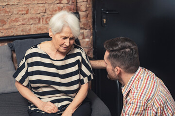 depressed elderly caucasian grandmother suffering from dementia sitting on a black couch and talking to her adult grandson. High quality photo