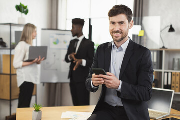 Caucasian businessman in suit using modern smartphone and looking at camera while sitting at office. Blur background of multiracial partner talking near flip chart.