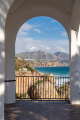 Beautiful landscape seen through an archway on Balcony of Europe , Nerja city, Malaga. View of La Chalaonda beach with Sierra Nevada mountains in background. Cloudy day. Vertical photography. - obrazy, fototapety, plakaty