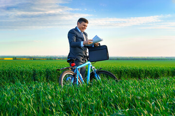 businessman dressed in a business suit, poses with bicycle in green grass field and reads documents, beautiful nature in spring, business concept