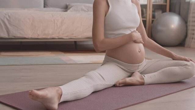Slowmo shot of young Caucasian pregnant woman in activewear doing stretching exercises sitting on yoga mat in bedroom