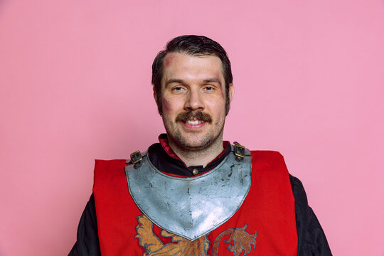 One young happy man, medieval warrior or knight wearing wearing armor clothing posing isolated over pink background. Comparison of eras, history, emotions
