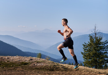 Side view of professional trail runner who taking part in summer marathon of running on mountain hills. Man with naked athletic body running against the backdrop of mountain panorama.
