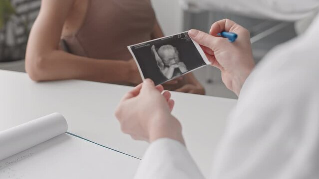 Slowmo closeup of unrecognizable female obstetrician holding ultrasound image of baby in hands while expectant mother stroking her belly in background having consultation in doctor office
