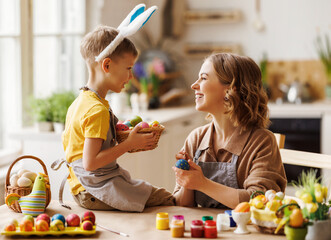 Loving young mother teaching happy little kid son to decorate Easter eggs while sitting in kitchen