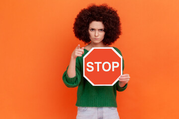 Fototapeta na wymiar Hey you, stop! Bossy woman with Afro hairstyle wearing green casual style sweater pointing finger to camera, showing red traffic stop sign in hands. Indoor studio shot isolated on orange background.