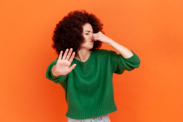 Fototapeta na wymiar Woman with Afro hairstyle grimacing with disgust, holding breath, pinching nose with fingers to avoid bad smell and showing stop gesture. Indoor studio shot isolated on orange background.
