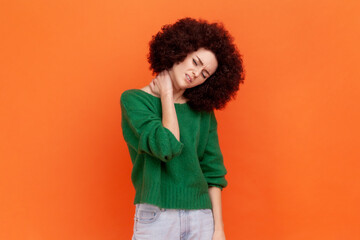 Health problems. Portrait of sick woman with Afro hairstyle wearing green casual style sweater standing and holding her painful neck. Indoor studio shot isolated on orange background.