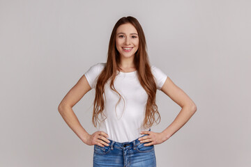 Portrait of woman standing with hands on hips and looking at camera with toothy smile, expressing positive emotions, wearing white T-shirt. Indoor studio shot isolated on gray background.