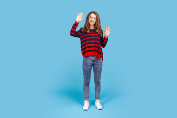 Hello! Full length portrait of attractive smiling woman wearing striped casual style sweater standing and waving hands, saying hi to friends. Indoor studio shot isolated on blue background.