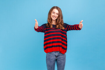 Positive friendly woman, giving free hugs with outstretched hands, welcoming inviting to embrace, wearing striped casual style sweater. Indoor studio shot isolated on blue background.