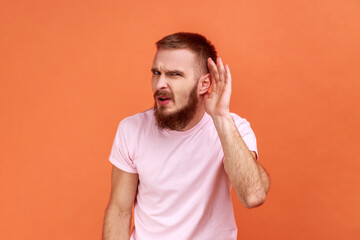 Fototapeta na wymiar Portrait of attentive handsome bearded man standing with hand on ear and want to hear something, wearing pink T-shirt. Indoor studio shot isolated on orange background.