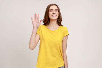 Hello! Portrait of charming teenager girl with brown hair, wearing casual style outfit standing, waving hand and saying Hi to camera with toothy smile. Indoor studio shot isolated on gray background.