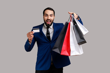 Bearded man holding and showing paper bags and credit card, contactless payments, easy shopping with debit card, wearing official style suit. Indoor studio shot isolated on gray background.