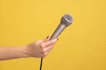 Profile side view closeup of human hand holding silver microphone for singing or reporting. Indoor studio shot isolated on yellow background.