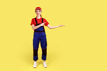 Full length portrait of happy worker woman pointing at copy space for advertisement on her palm, looking at camera with smile, wears overalls and cap. Indoor studio shot isolated on yellow background.