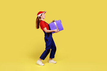 Side view portrait of courier woman carrying heavy presents boxes for holidays, door-to-door delivery, wearing overalls and red cap. Indoor studio shot isolated on yellow background.