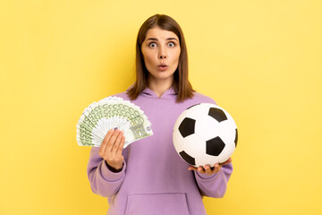 Portrait of woman with open mouth holding soccer ball and fan of euro, amazed of football championship, betting, wearing purple hoodie. Indoor studio shot isolated on yellow background.