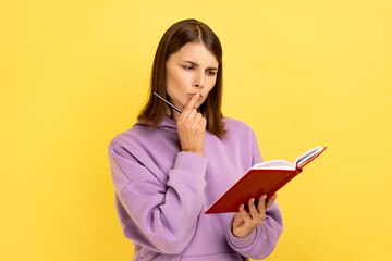 Thoughtful woman standing with paper notebook and pen, looking away with pensive expression, making to do list, wearing purple hoodie. Indoor studio shot isolated on yellow background.