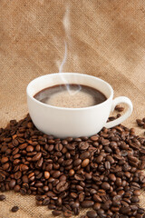 Cup with coffee and coffee beans and steam over cup on burlap background