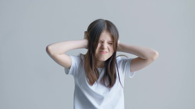 A blonde teenager girl in a white T-shirt covered her ears with her hands, is unhappy and shakes her head against the background of a white wall