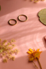 Gold wedding rings and various pressed flowers on pastel pink background, illuminated by sunlight. Selective focus.