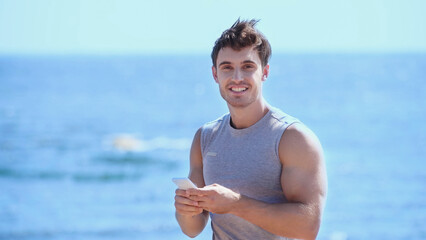 Positive sportsman using smartphone and looking at camera near blurred sea