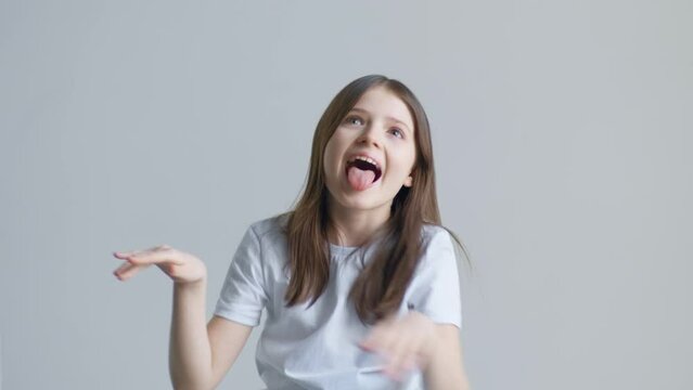 A blonde teenager girl in a white T-shirt grimaces and shows her tongue against a white wall