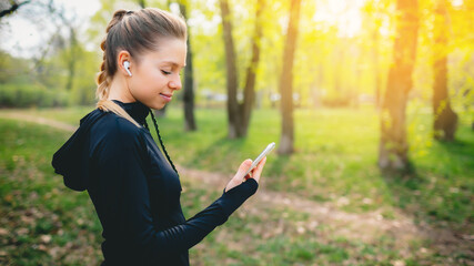 Young smiling girl making sport and running in the park using her phone to listen the music with wireless headphones on sunset in the city watching the screen