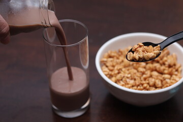 Chocolate milk is pouring into a glass. Ready cereal in a white cup.