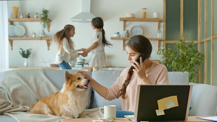 A Busy Mother Trying to Work Remotely at Home. On The Background Her Daughters are Running Around The Table, While Their Mother Working With a Laptop and Doing Business Conversation on the Phone.
