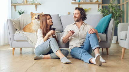 Happy Young Married Couple Sitting on the Floor Near a Couch and Clinking Glasses With Wine,...
