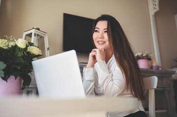 Young Beautiful Asian business woman working online with laptop computer placed at the table at home office. Smiling lady freelancer have remote work sitting in living room with white roses flowers
