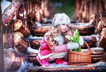 Baby in the winter forest. Girl on a wooden bridge. Baby in winter coat. Girl in early spring. Beautiful girl with long hair in a hat. Girl in a sweater. containing tulips. girl smelling flowers