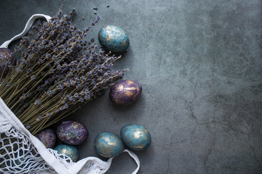 Easter Card With A Copy Of The Place For The Text. Purple, Blue And Golden Eggs In A String Bag On A Dark Background. The Trend Of 2022 Purple Shade Is Very Peri. Natural Dye Karkade Tea. Top View.