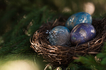Happy Easter 2022. A postcard with a copy of the place for the text. Purple, blue and golden eggs in a nest on a natural forest background. Top view.