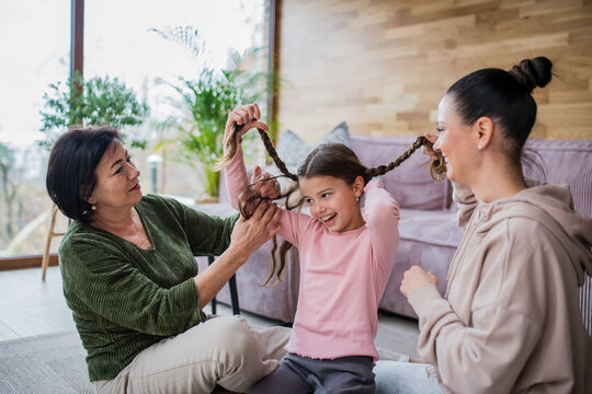 Happy little girl sitting on floor, her mother and grandmother making her plaits at home.