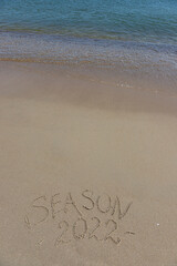 The inscription season 2022 on the sand by the water and the rising wave, seashore beach vacation by the sea