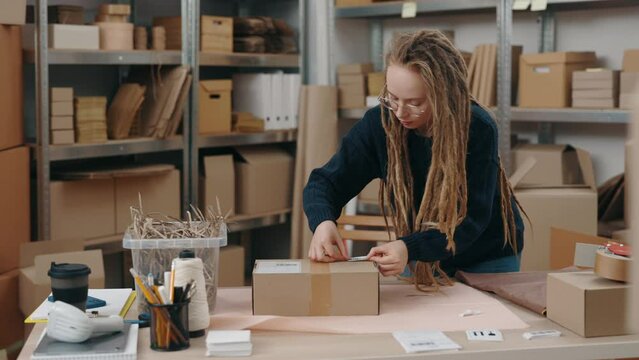 At my workplace. Calm caucasian woman with dreadlocks wearing glasses putting sticker with barcode at the cupboard box while working at the warehouse. Small business concept.